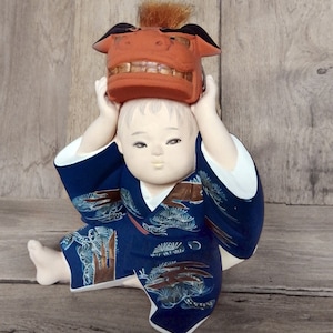 May include: A ceramic doll wearing a blue kimono with a gold and white pine tree pattern. The doll is holding a red and brown lion mask above its head.
