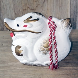 May include: A white ceramic Japanese guardian dog figurine with gold accents and a red and white braided cord. The figurine has black eyes and red accents on its face. The dog is sitting down.