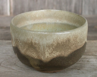 May include: A ceramic tea bowl with a light brown glaze and a darker brown rim. The bowl has a speckled appearance and a slightly uneven surface.