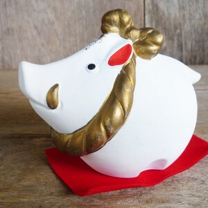 May include: White ceramic boar figurine with gold rope detail and red accents. The boar is sitting on a red fabric mat.