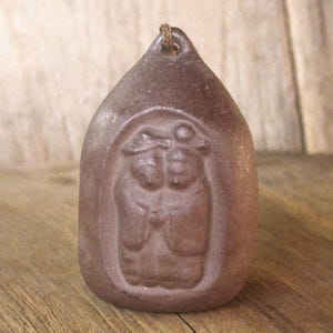 May include: A brown, teardrop-shaped ceramic pendant with a carved relief of two figures. The pendant has a loop at the top for hanging and a textured surface. The figures are stylized and appear to be wearing hats.