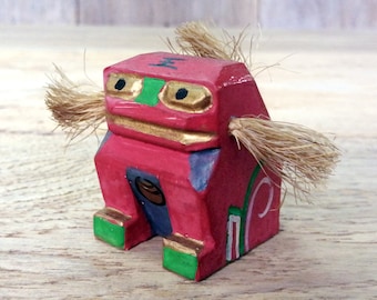 May include: A small, red wooden figurine with a gold and green painted design. The figurine has two long, brown, furry ears and a small, round, brown nose. The figurine is sitting on a wooden surface.