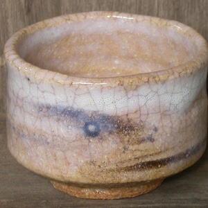 Può includere: Una piccola tazza da tè in ceramica bianca con una glassa craquelé e accenti marroni e blu. La tazza ha una forma arrotondata e un bordo largo.