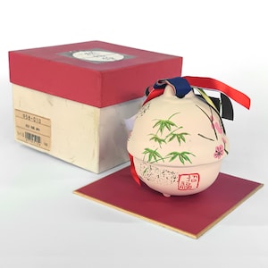 Könnte beinhalten: Ein rosafarbener Keramik-Anhänger mit einem Bambus- und Kirschblüten-Design. Der Anhänger ist mit bunten Bändern gebunden und hat ein rotes Siegel mit dem chinesischen Schriftzeichen für "Glück" auf der Unterseite. Der Anhänger befindet sich in einer roten und weißen Schachtel.