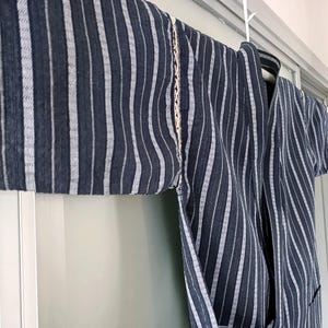 Peut inclure: Un kimono à rayures bleu foncé et blanches est suspendu à un cintre blanc. Le tissu présente des rayures verticales de largeurs variables. Une garniture décorative crème longe la couture de l'épaule. Le kimono est fait d'un tissu léger.