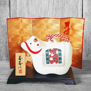 Puede incluir: Una figurita de vaca de cerámica blanca con diseños florales rojos y verdes. La vaca está sentada sobre una base de madera y tiene una cuerda roja y blanca atada a su cabeza. La vaca está delante de una pantalla dorada con el carácter japonés para "primavera" escrito en ella.