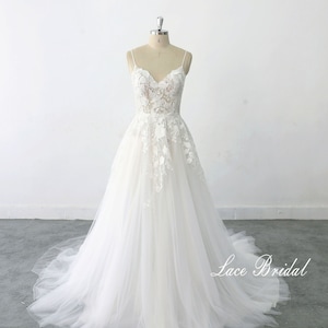 May include: A white lace wedding dress with a v-neckline and spaghetti straps. The dress has a tulle skirt and is adorned with floral lace appliques. The dress is displayed on a mannequin against a white background. The text 'Lace Bridal' is visible at the bottom of the image.