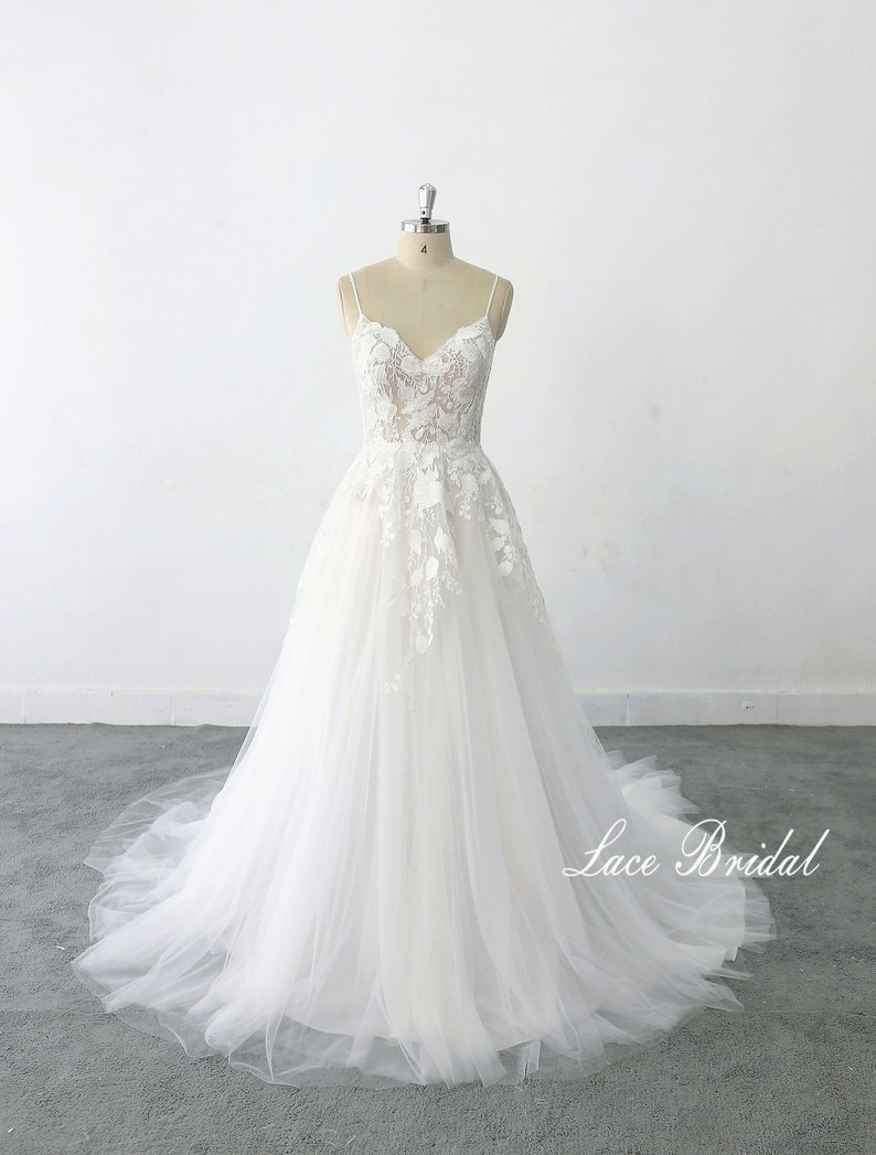 May include: A white lace wedding dress with a v-neckline and spaghetti straps. The dress has a tulle skirt and is adorned with floral lace appliques. The dress is displayed on a mannequin against a white background. The text 'Lace Bridal' is visible at the bottom of the image.