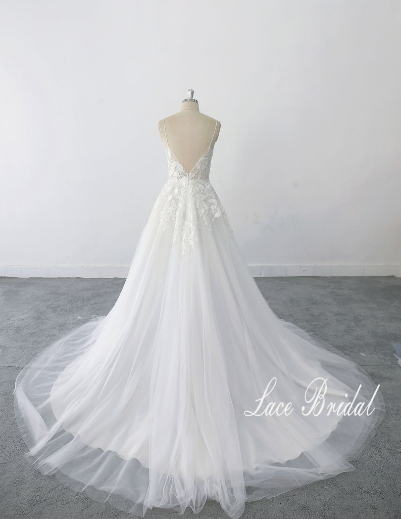 May include: A white wedding dress with a lace bodice and a tulle skirt. The dress has a deep V-neckline and a low back. The dress is on a mannequin and the skirt is flowing out behind it. The text 'Lace Bridal' is visible in the bottom right corner of the image.
