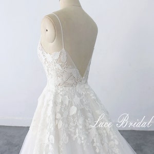 May include: A white lace wedding dress with a v-neckline and spaghetti straps. The dress has a fitted bodice and a flowing skirt. The skirt is decorated with floral lace appliques. The dress is displayed on a mannequin against a white background. The text 'Lace Bridal' is visible on the bottom right of the image.
