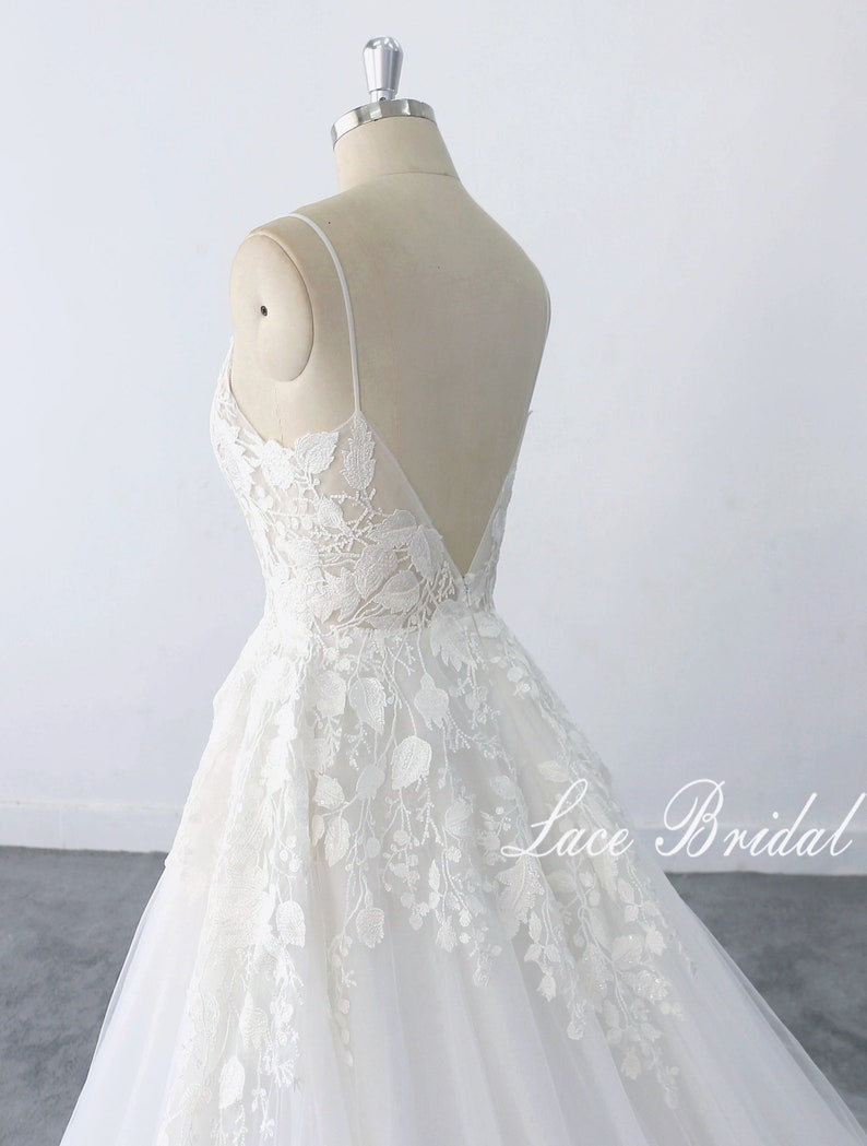 May include: A white lace wedding dress with a v-neckline and spaghetti straps. The dress has a fitted bodice and a flowing skirt. The skirt is decorated with floral lace appliques. The dress is displayed on a mannequin against a white background. The text 'Lace Bridal' is visible on the bottom right of the image.