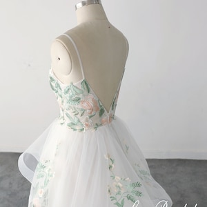 May include: A white tulle wedding dress with a fitted bodice and a layered skirt. The dress features delicate floral embroidery in shades of green and pink. The back of the dress has a low, V-shaped neckline and thin straps.