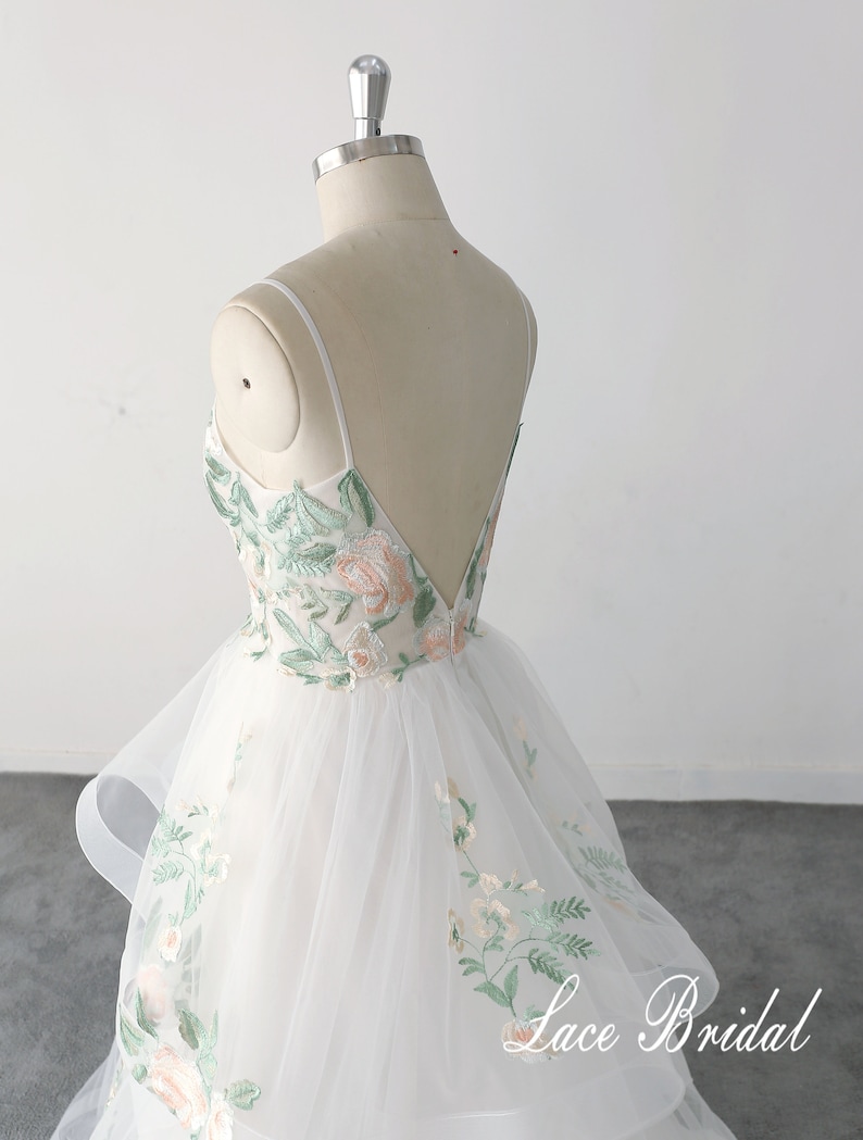 May include: A white tulle wedding dress with a fitted bodice and a layered skirt. The dress features delicate floral embroidery in shades of green and pink. The back of the dress has a low, V-shaped neckline and thin straps.
