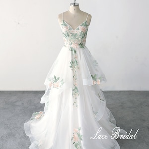 May include: A white tulle wedding dress with a V-neckline and spaghetti straps. The dress is adorned with delicate floral embroidery in shades of green and pink. The skirt features multiple layers of tulle, creating a flowing and romantic silhouette. The dress is displayed on a mannequin and the text "Lace Bridal" is visible at the bottom of the image.