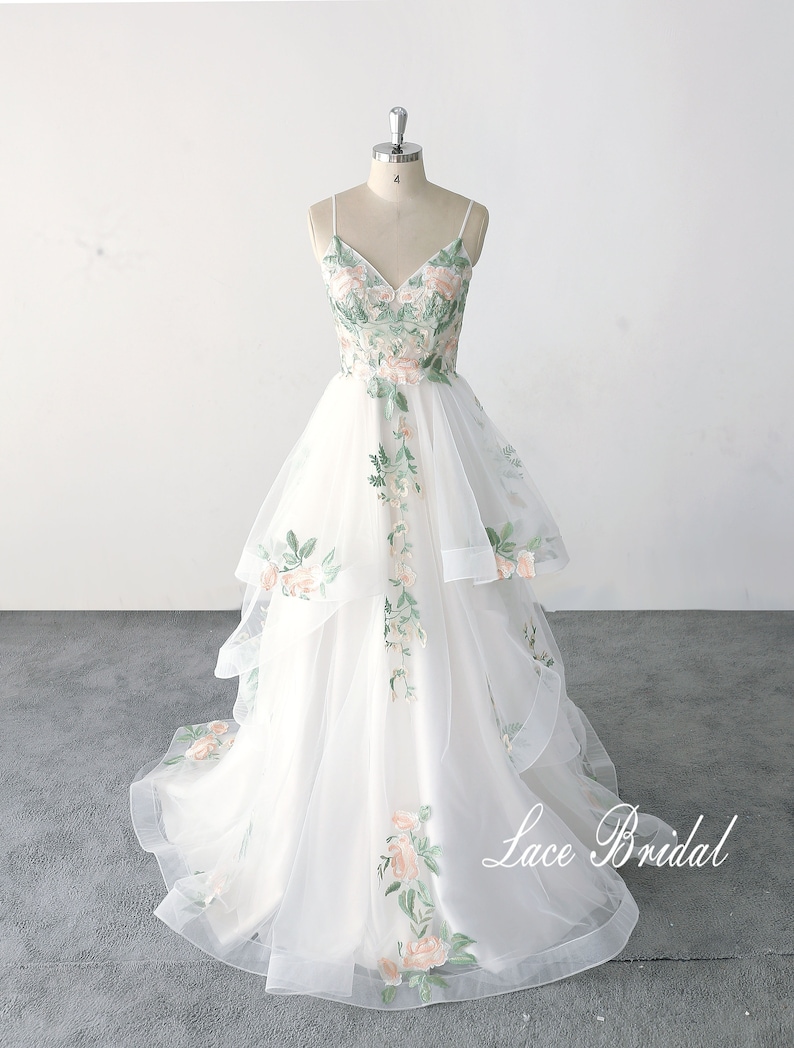 May include: A white tulle wedding dress with a V-neckline and spaghetti straps. The dress is adorned with delicate floral embroidery in shades of green and pink. The skirt features multiple layers of tulle, creating a flowing and romantic silhouette. The dress is displayed on a mannequin and the text "Lace Bridal" is visible at the bottom of the image.