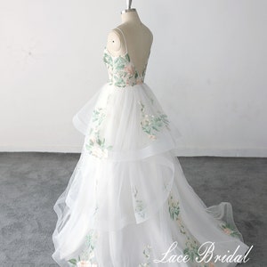 May include: A white tulle wedding dress with a floral embroidery design. The dress has a sweetheart neckline and a layered skirt. The dress is on a mannequin and the text "Lace Bridal" is visible on the bottom of the skirt.