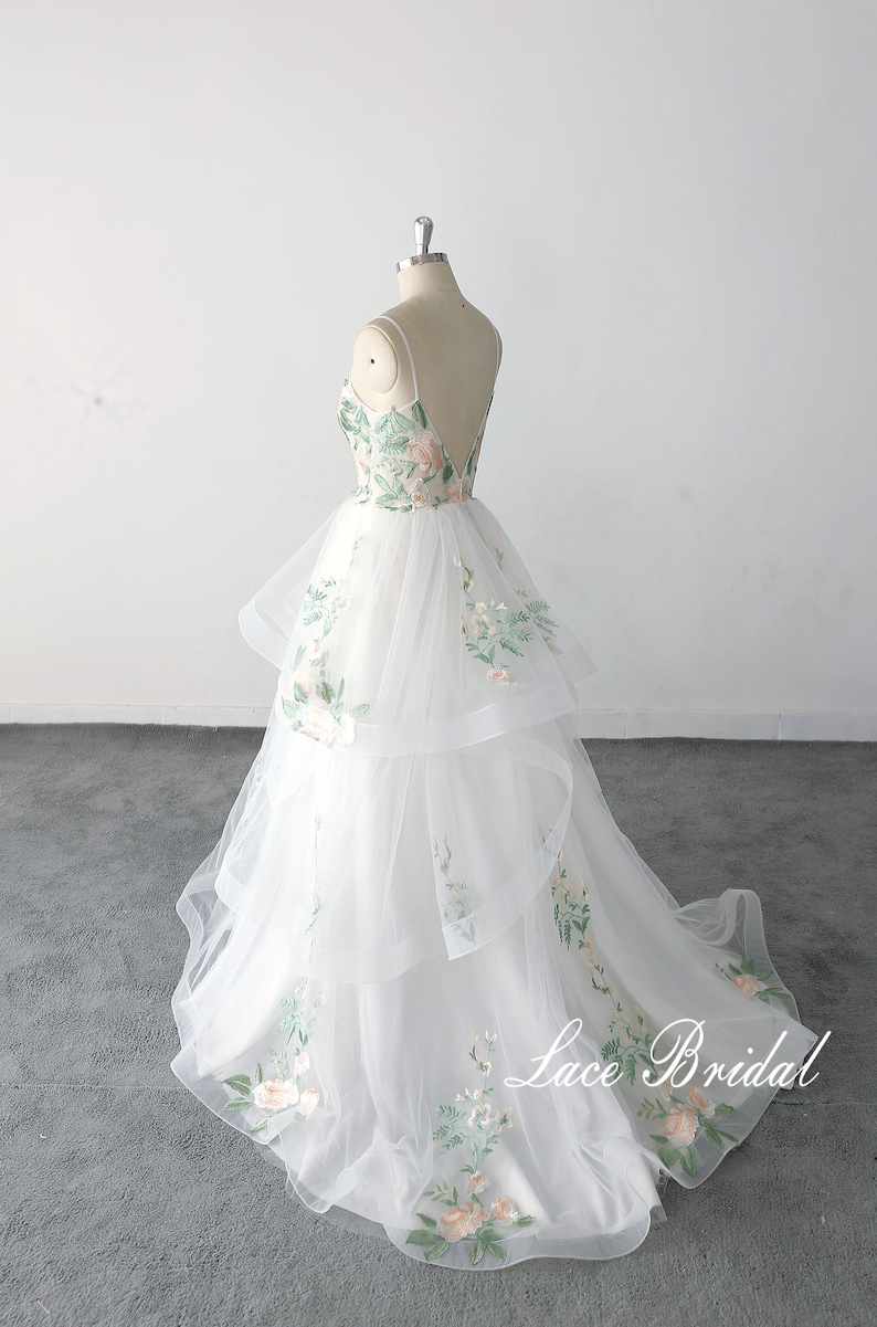 May include: A white tulle wedding dress with a floral embroidery design. The dress has a sweetheart neckline and a layered skirt. The dress is on a mannequin and the text "Lace Bridal" is visible on the bottom of the skirt.