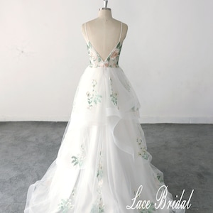 May include: A white tulle wedding dress with a floral embroidery design. The dress has a V-shaped back and a layered skirt. The dress is on a mannequin and the text "Lace Bridal" is visible at the bottom of the image.