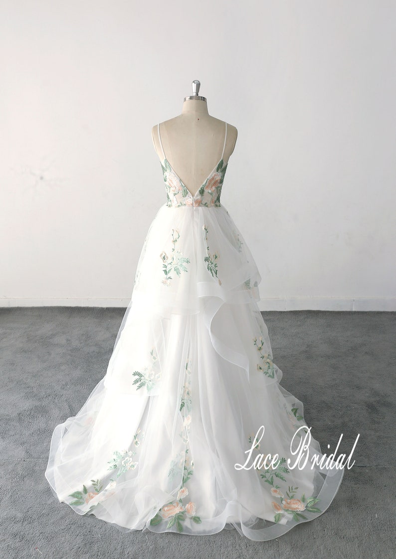 May include: A white tulle wedding dress with a floral embroidery design. The dress has a V-shaped back and a layered skirt. The dress is on a mannequin and the text "Lace Bridal" is visible at the bottom of the image.