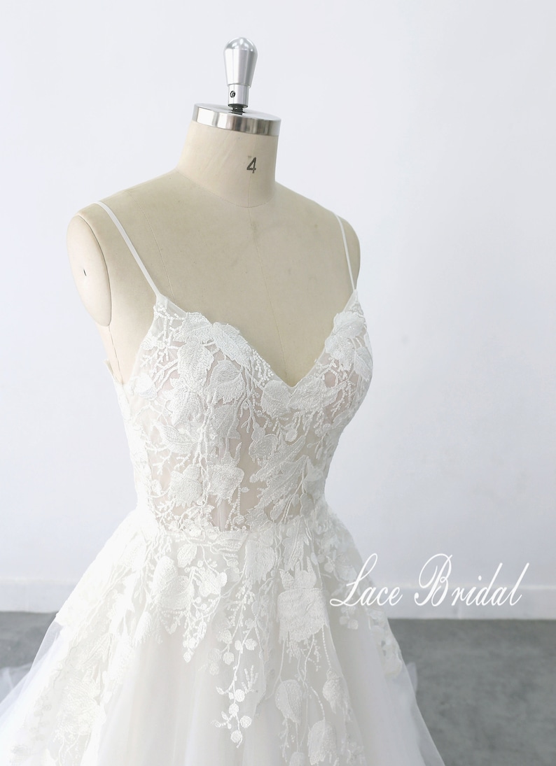 May include: A white lace wedding dress with a sweetheart neckline and spaghetti straps. The dress has a full skirt and is adorned with delicate floral lace. The dress is displayed on a mannequin against a white background. The text 'Lace Bridal' is visible in the bottom right corner of the image.