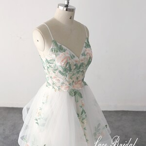 May include: A white tulle wedding dress with a V-neckline and spaghetti straps. The dress is embroidered with green leaves and pink flowers. The skirt is layered and has a flowing silhouette. The dress is on a white mannequin. The text "Lace Bridal" is visible in the bottom right corner of the image.