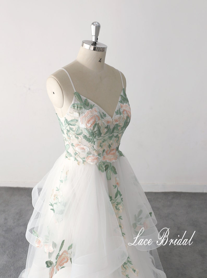 May include: A white tulle wedding dress with a V-neckline and spaghetti straps. The dress is embroidered with green leaves and pink flowers. The skirt is layered and has a flowing silhouette. The dress is on a white mannequin. The text "Lace Bridal" is visible in the bottom right corner of the image.