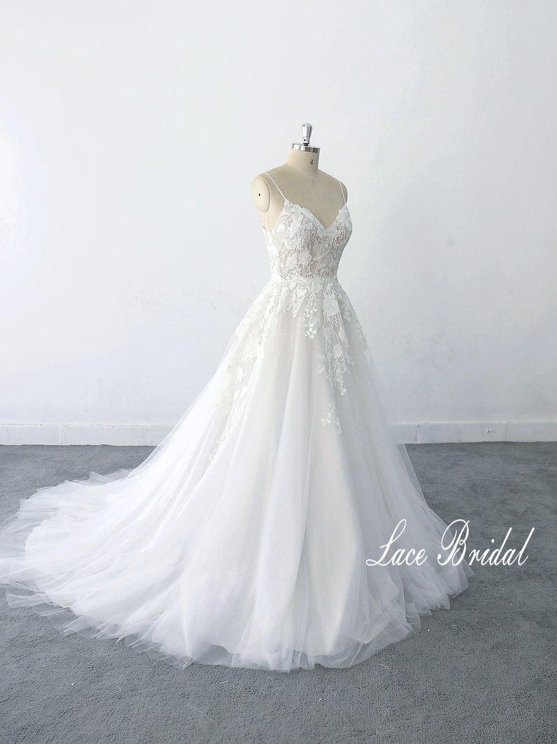 May include: A white lace wedding dress with a v-neckline and spaghetti straps. The dress has a tulle skirt and is adorned with floral lace appliques. The dress is displayed on a mannequin against a white background. The text 'Lace Bridal' is visible in the bottom right corner of the image.