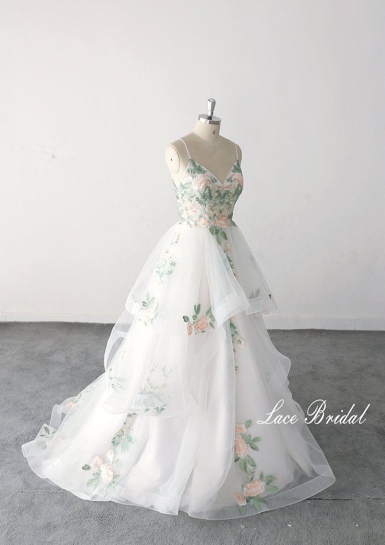 May include: A white tulle wedding dress with a floral embroidery design. The dress has a V-neckline and a layered skirt. The dress is on a mannequin and the background is white.