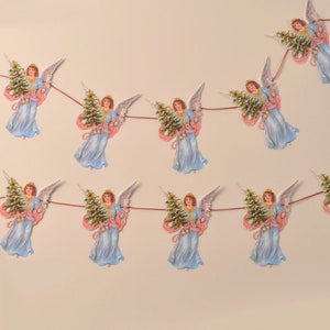 May include: A festive garland featuring paper cutouts of angels holding Christmas trees. The angels wear light blue gowns with pink accents and gold halos. The garland is strung on a thin red cord, perfect for holiday decor.