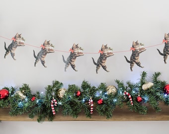 May include: A festive garland featuring a repeating series of tabby cats standing on their hind legs, each wearing a red bow and carrying a small Christmas pudding. The cats are strung together on a thin red cord, positioned above a green pine garland.