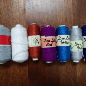 May include: Eight spools of yarn in various colors, including gray, white, orange, purple, blue, and two shades of blue. The spools are labeled with the text "Dee Lite Yellow" and "Dee Lite Red".