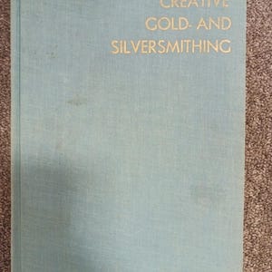 May include: A light blue book titled "CREATIVE GOLD- AND SILVERSMITHING" in gold lettering. The book has a textured cover and is slightly worn. The book is lying on a brown surface.