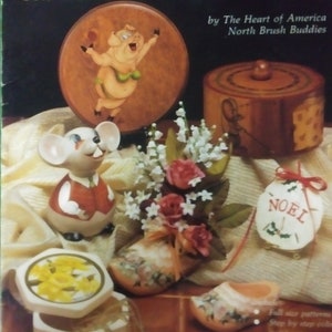 May include: A book titled "Sharing Ideas with America's Tole and Decorative Painter's" by The Heart of America North Brush Buddies. The book cover features a variety of painted items, including a pig, a mouse, a flower arrangement, a wooden box, and a pair of wooden shoes.