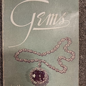 Op de afbeelding: Een vintage boekomslag met de titel "Gems" met een lichtblauwe achtergrond. De omslag toont een diamanten ketting met een grote, donkerpaarse hanger. De tekst "in the SMITHSONIAN INSTITUTION" staat onderaan. Een prijskaartje van $4 staat rechtsboven.