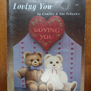 May include: A book cover with the title "Loving You" by Camille & Sue Scheewe. The cover features a red heart with the words "Loving You" inside. Two teddy bears are sitting on a purple bench with a heart pattern. The book is for a craft project.