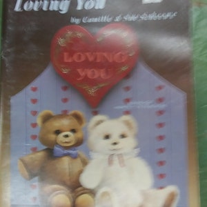 May include: A book titled "Loving You" by Camille & Sue Scheene with a red heart illustration that says "Loving You" and two teddy bears, one brown and one white, sitting on a purple background with small red hearts.