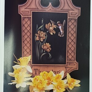 May include: A wooden sign with a carved frame and a black background. The sign features a hummingbird in flight, a floral design with yellow daffodils, and the text "Thornton".