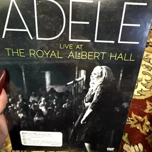 May include: DVD of Adele's "Live at The Royal Albert Hall" concert. The cover features the artist performing on stage with a microphone, in front of a crowd. The title "ADELE" is in large white letters, with the venue name below.