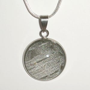 May include: A circular silver-toned pendant featuring a textured, dark gray, and metallic-flecked inlay. The pendant is suspended from a delicate silver-toned chain, showcasing a polished metal frame that secures the circular centerpiece in place.