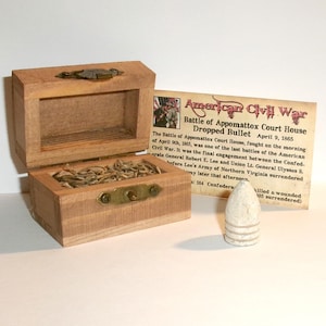 May include: A small wooden box with a brass latch, containing fibrous material, accompanied by a conical lead bullet and a rectangular information card titled American Civil War Battle of Appomattox Court House Dropped Bullet April 9, 1865.