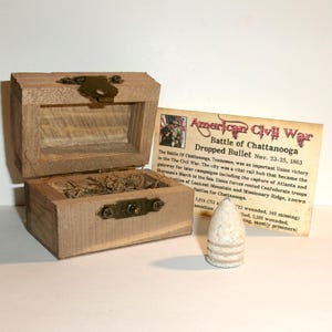 May include: A small, light-brown wooden box with a brass-colored latch, containing dried grass. Beside it sits a conical, off-white metal bullet and a rectangular information card titled American Civil War Battle of Chattanooga Dropped Bullet.