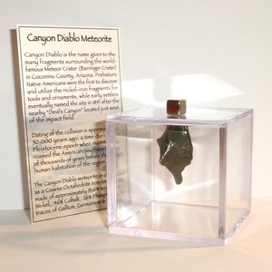 May include: A Canyon Diablo meteorite fragment suspended inside a clear acrylic cube display case. A small metallic cube sits on top of the lid. Beside the display is an informational card titled Canyon Diablo Meteorite with text.