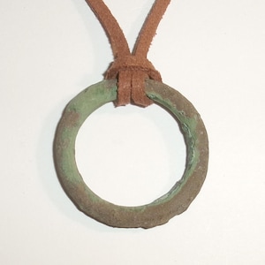 May include: A circular pendant featuring a weathered, textured metal ring with a distinct green patina and brown oxidation. The ring is suspended from a simple brown leather cord that is looped through the top of the metal circle in a knot.