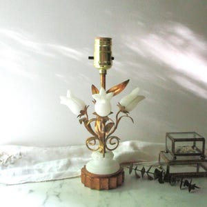 May include: A table lamp featuring a brass-toned metal base and stem with three white frosted glass tulip-shaped shades. The lamp has a central brass socket and a circular, fluted base. The metal structure includes decorative leaf and vine accents.