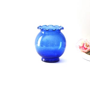 May include: A small, round, cobalt blue glass vase with a ruffled rim. The vase has a bulbous body and a short, flared base. The glass has a slightly textured appearance. The vase is likely used for displaying flowers or as a decorative item.