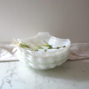 May include: A set of three white milk glass bowls with a scalloped edge. The bowls are stacked on top of each other and a sprig of white flowers is resting on top of the bowls.