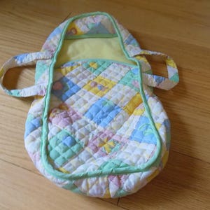 May include: A quilted baby carrier in pastel colors, including yellow, blue, pink, and green. The carrier has a rounded shape with a handle and a light green trim. The interior is a solid yellow.