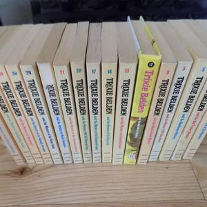 May include: A collection of 19 paperback books with yellow covers and the title "Trixie Belden" printed on the spines. The books are arranged in a row, with the spines facing the viewer. The books are numbered 1 through 21, with the exception of number 10, which is missing.