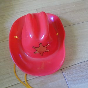 May include: A bright red plastic cowboy hat featuring a moulded crown and a wide brim. The front of the hat displays a gold-coloured six-pointed sheriff star emblem. A thin yellow cord is attached to the sides of the hat for securing it in place.