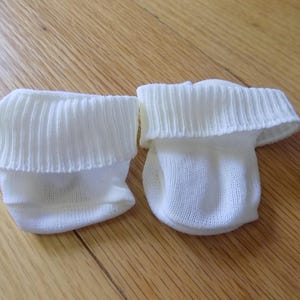 May include: A pair of plain white baby socks featuring a soft, knitted texture and a wide, ribbed cuff at the top. The socks are constructed from a lightweight fabric and are designed with a simple, rounded shape to fit comfortably on small feet.